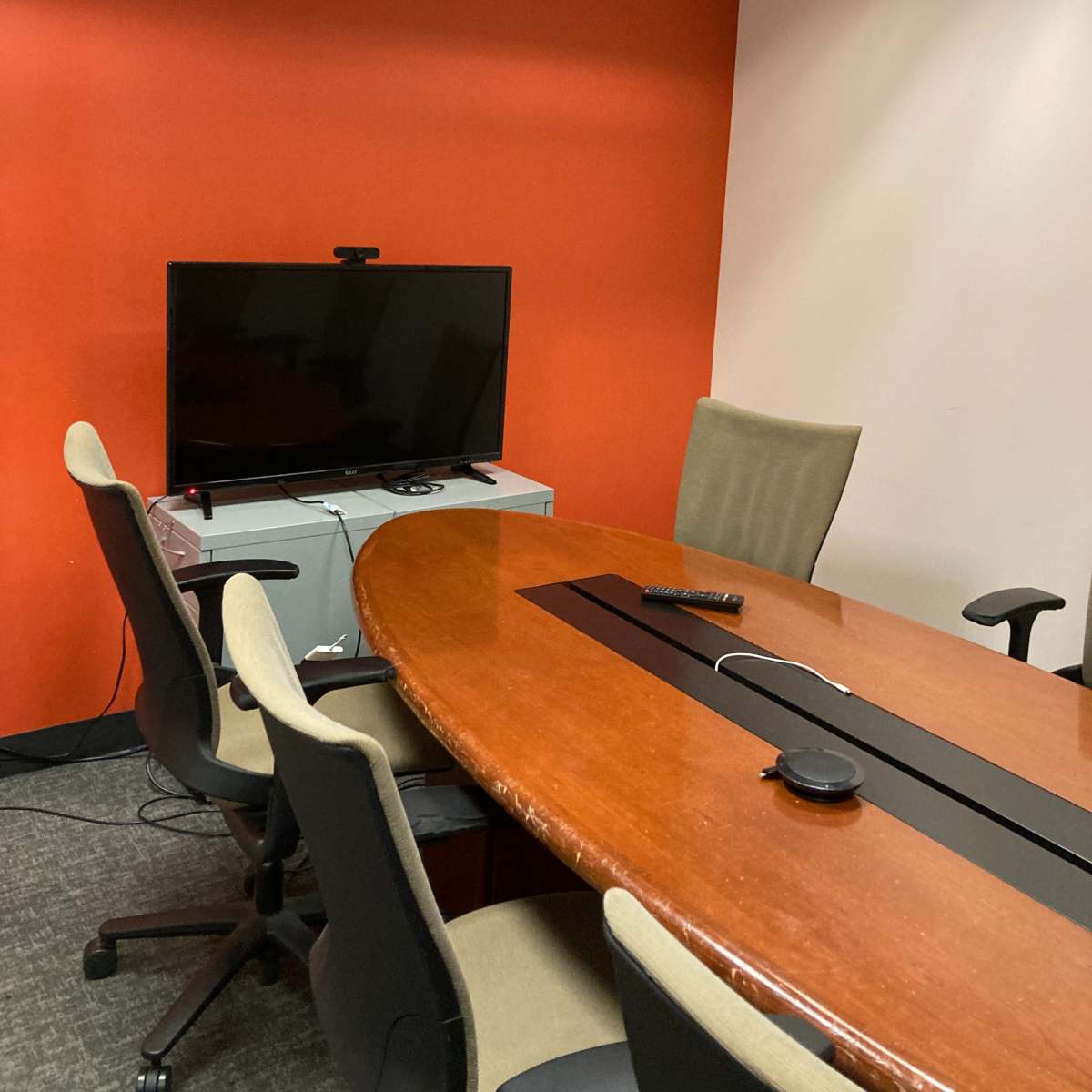 A small conference room with a round wooden table, several chairs, a television mounted on the wall, and an orange accent wall.
