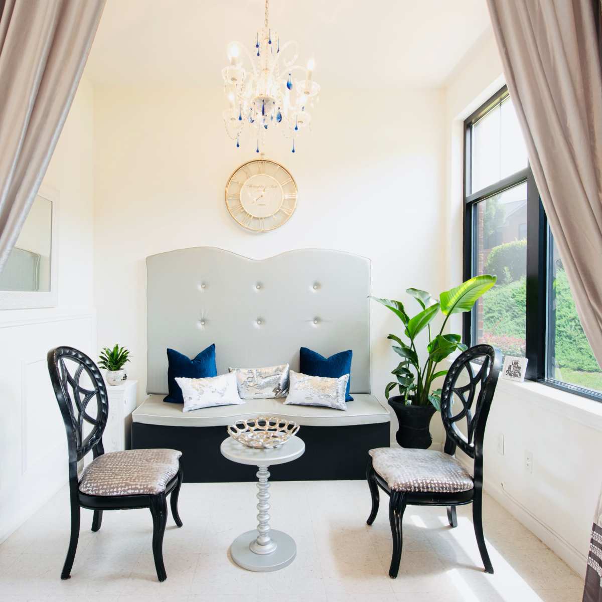 A small sitting area with a tufted gray bench, two black chairs, a round table, and a large window with nature visible outside.