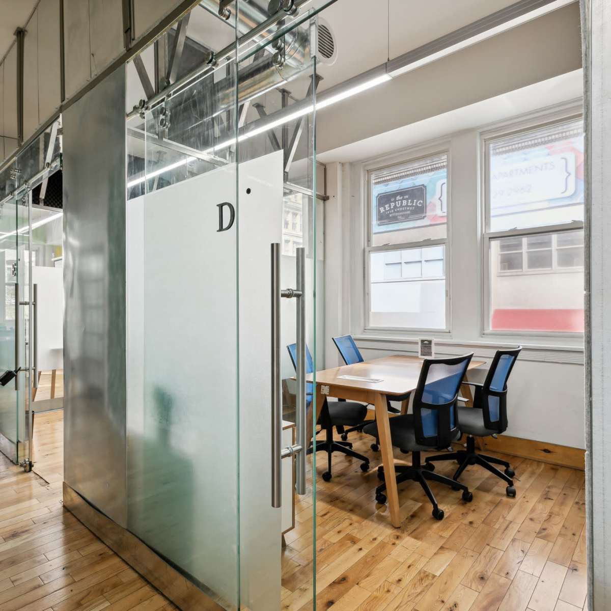 A modern office space featuring glass meeting rooms and wooden flooring, with several desks and chairs visible.