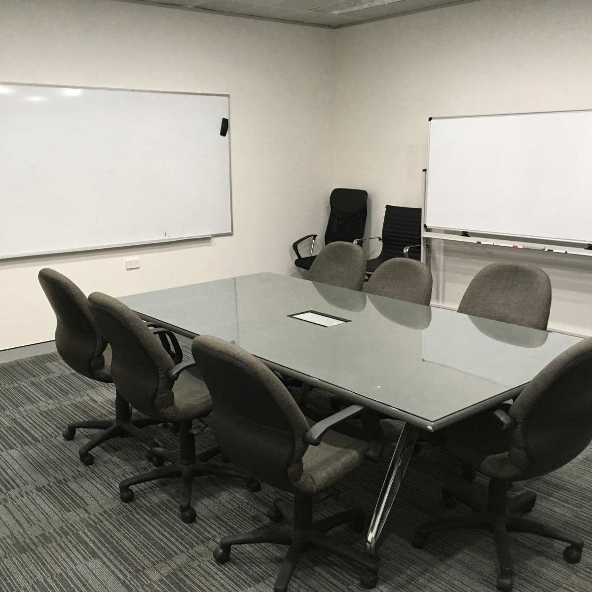 A glass-topped conference table surrounded by several office chairs in a meeting room with whiteboards on the walls.