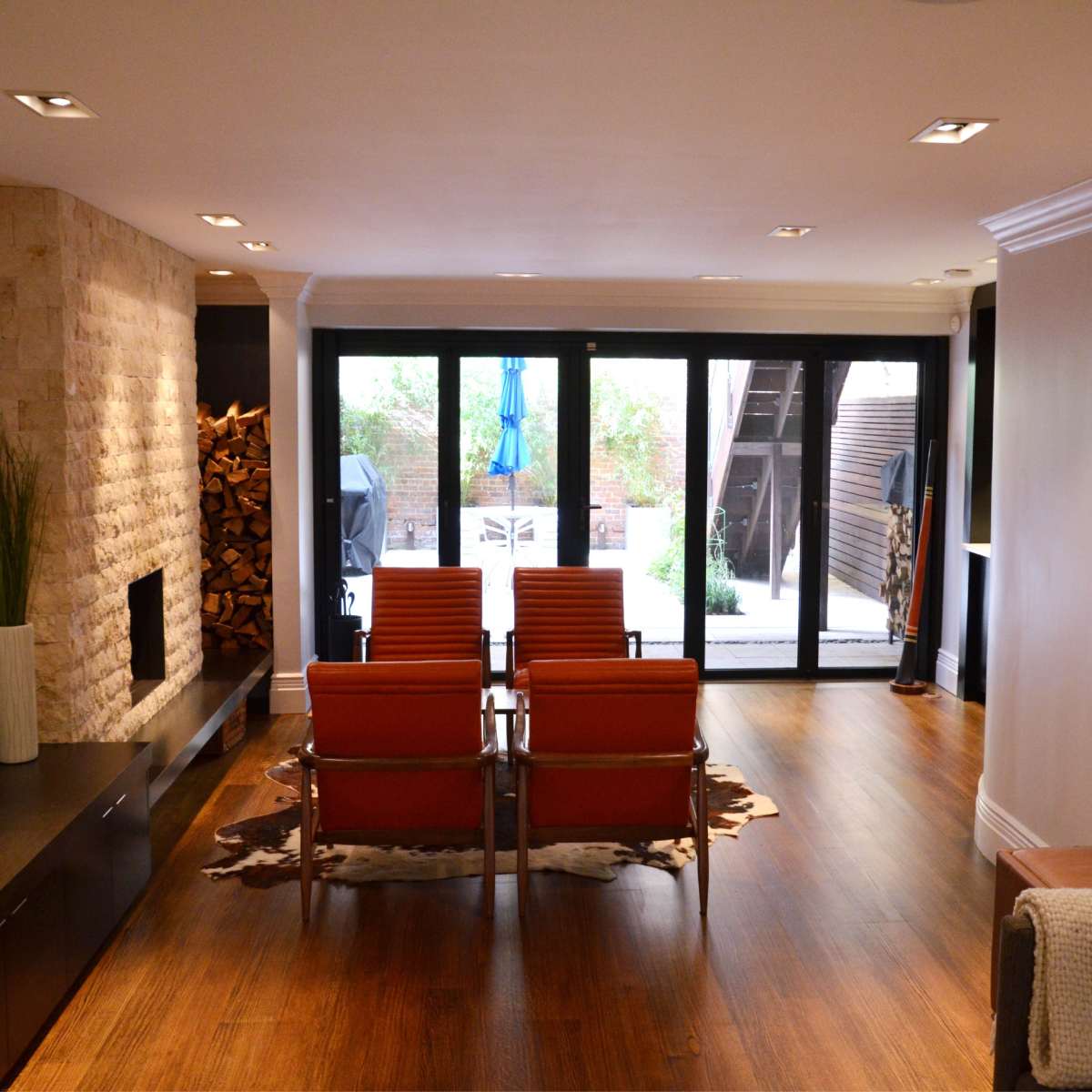 A modern living room with a stone fireplace, wooden flooring, and orange chairs facing large glass doors that open to a backyard.