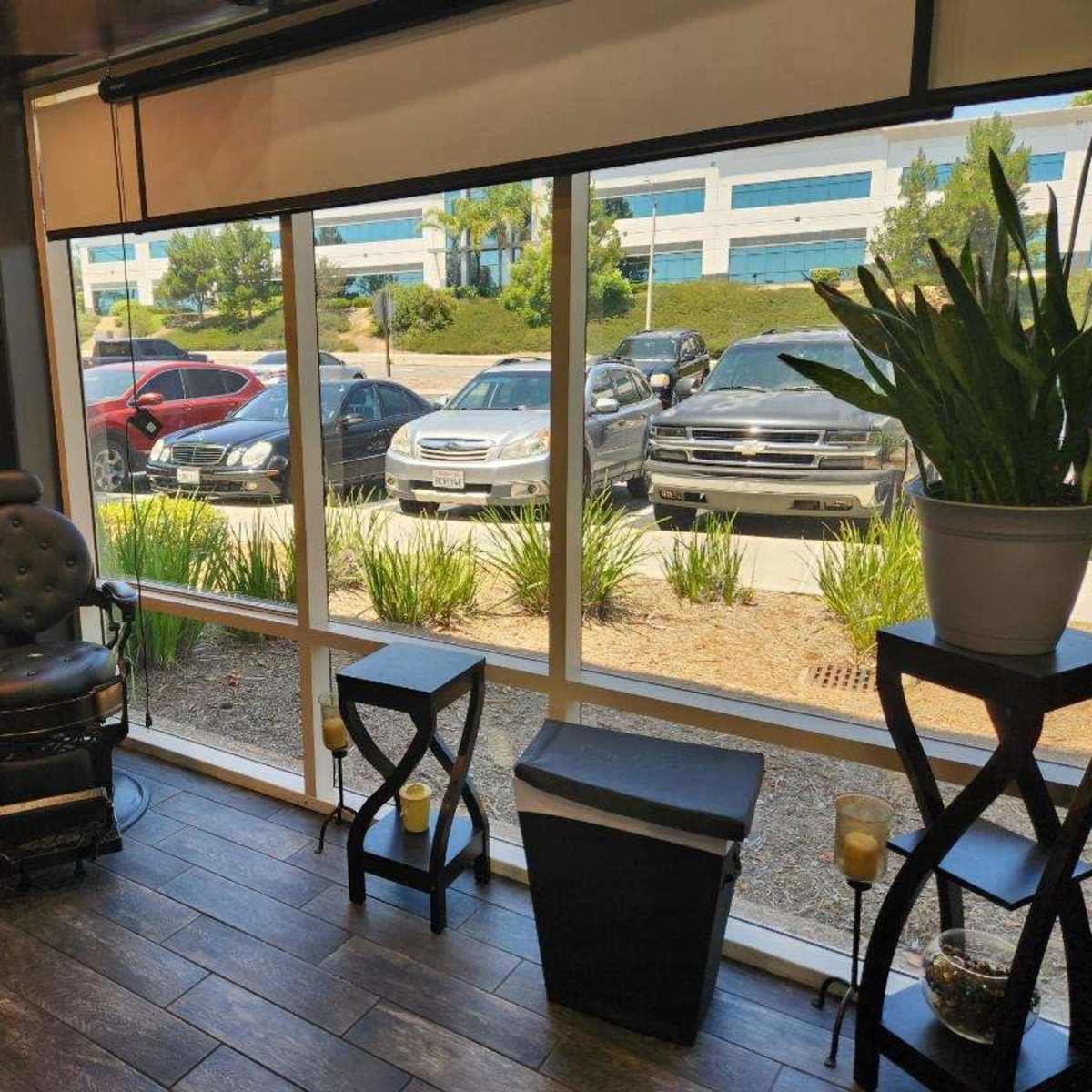 A barber shop interior with a black barber chair next to large windows overlooking parked cars and greenery outside.