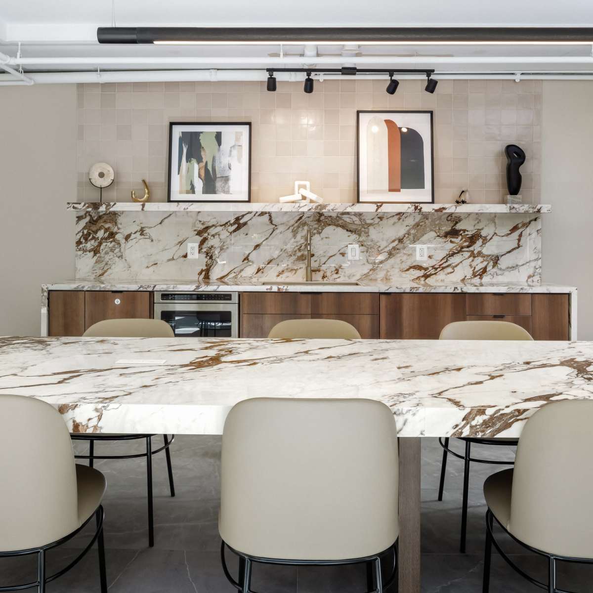 A modern kitchen features a large marble countertop with beige chairs and a stone backsplash displaying framed artwork.