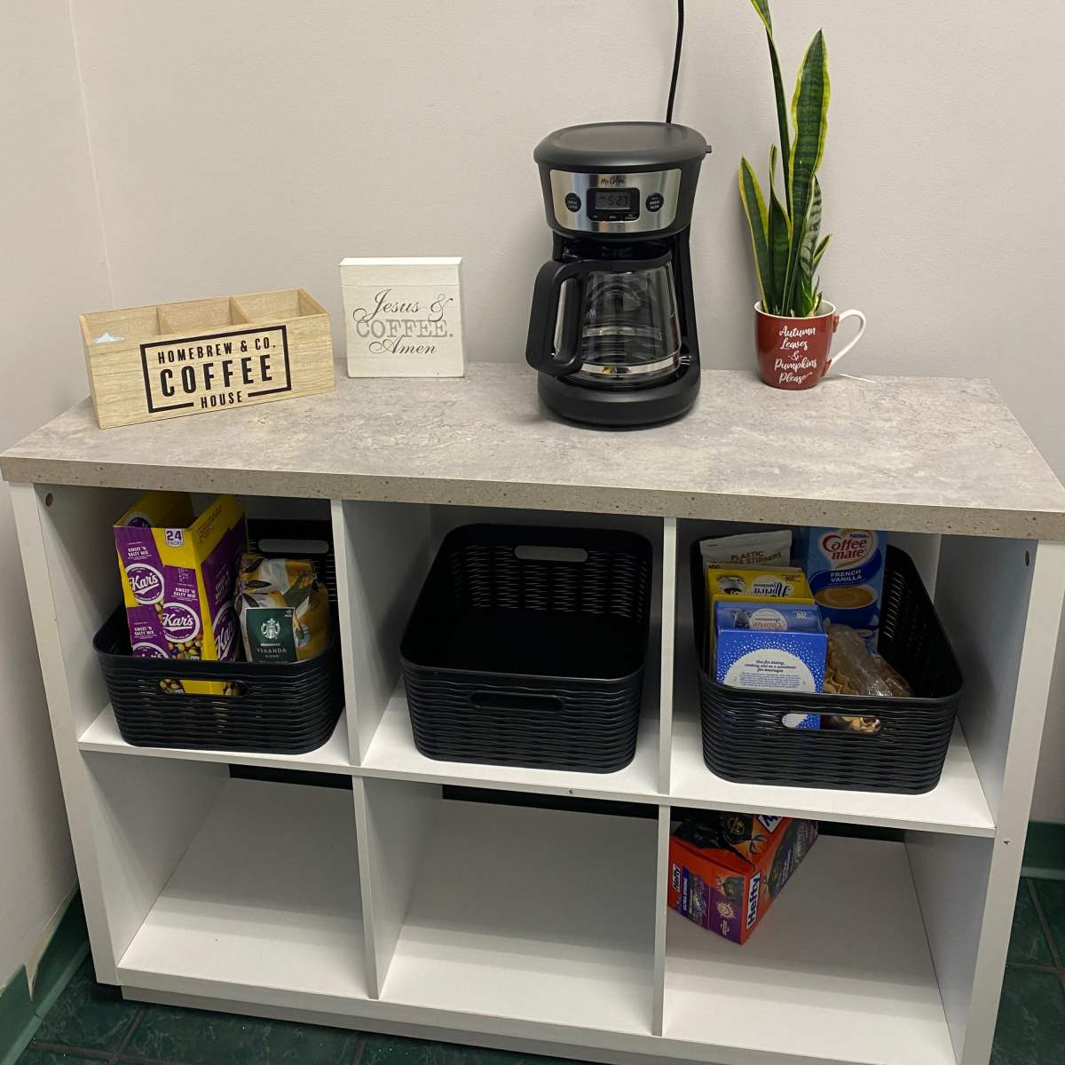 A small coffee station featuring a coffee maker, decorative signs, and organized storage baskets on a white shelf.