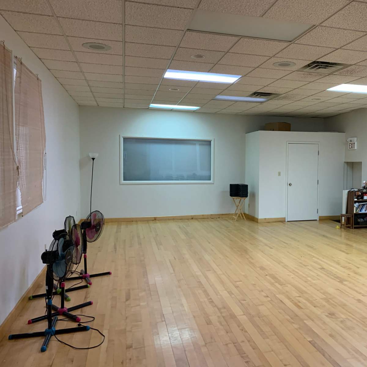 An empty room with wooden floors, a large window, and two standing fans positioned against one wall.