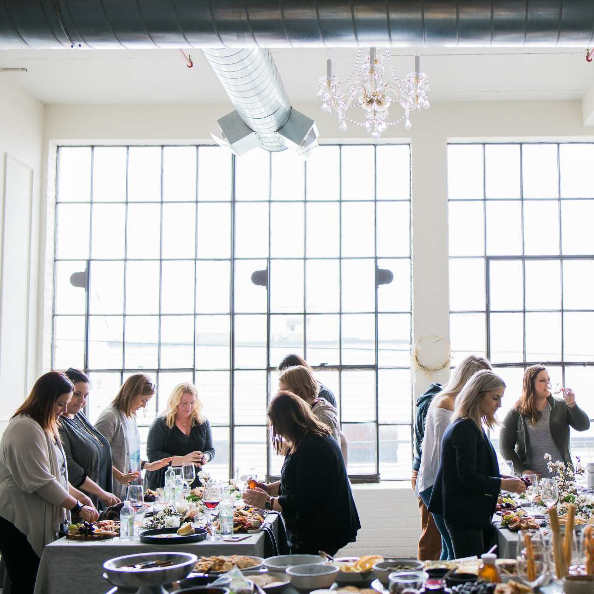 A group of women gather around a dining table, enjoying food and drinks in a spacious room with large windows.