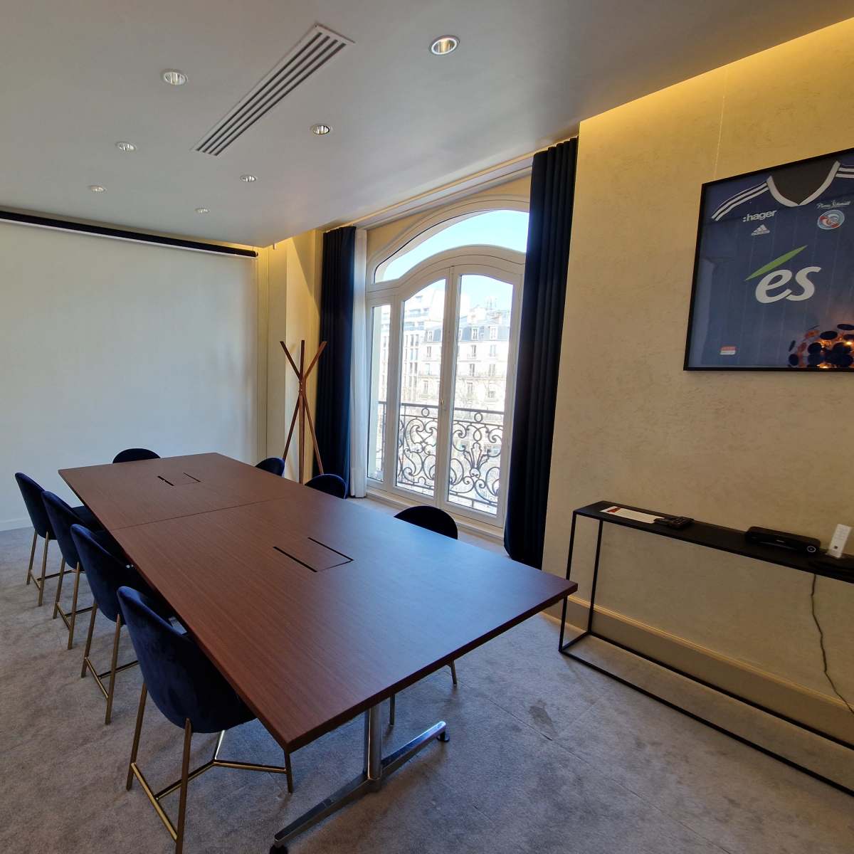 A modern meeting room features a long wooden table surrounded by blue chairs, with a large window offering a view of the city outside.