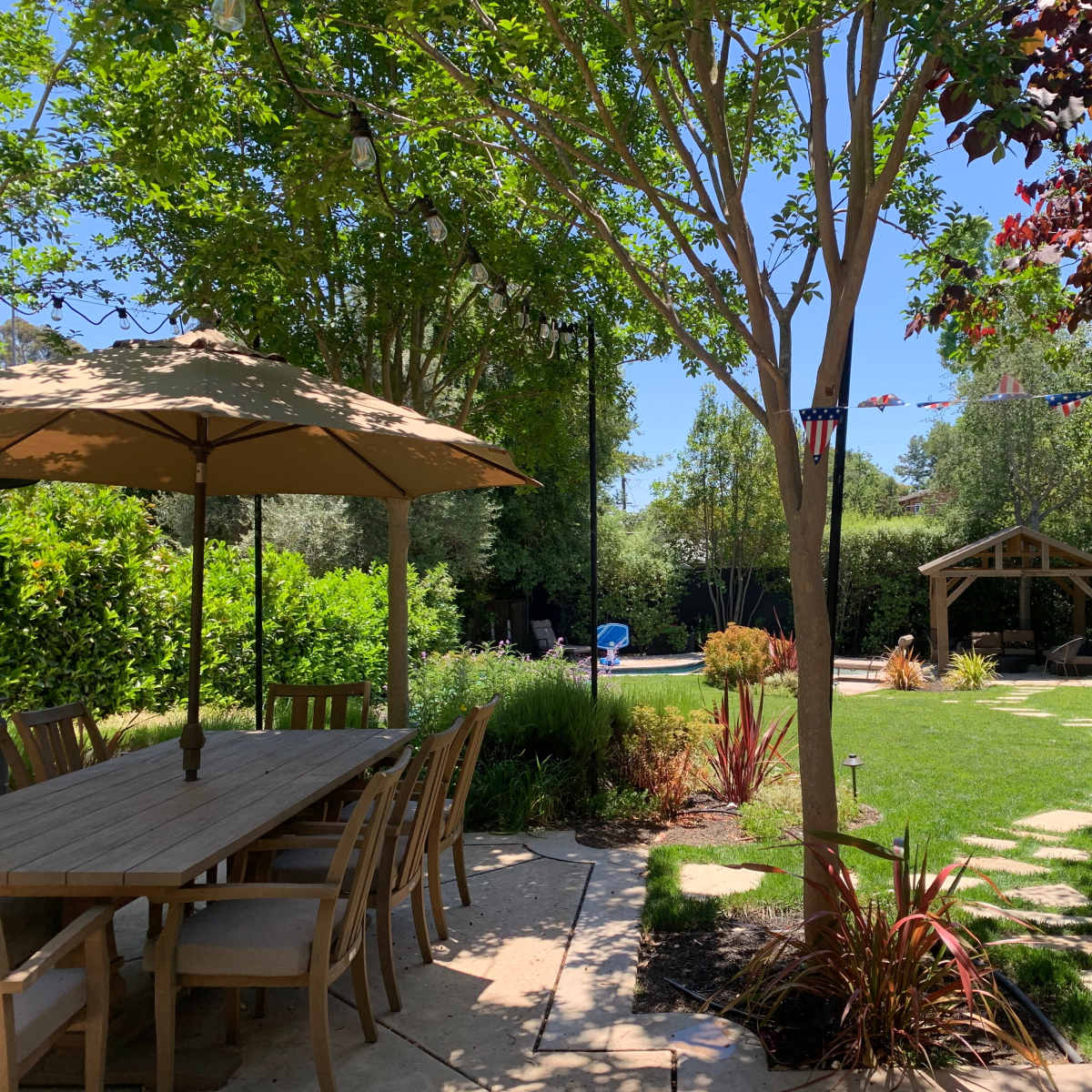 A patio area with a wooden dining table and umbrella overlooks a landscaped garden featuring a lawn, stone pathway, and a gazebo.