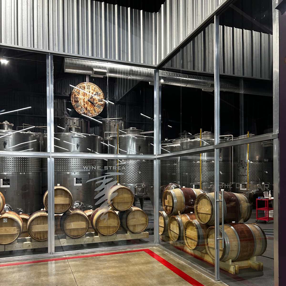 A glass-enclosed room with stacked wine barrels and stainless steel fermentation tanks in the background.