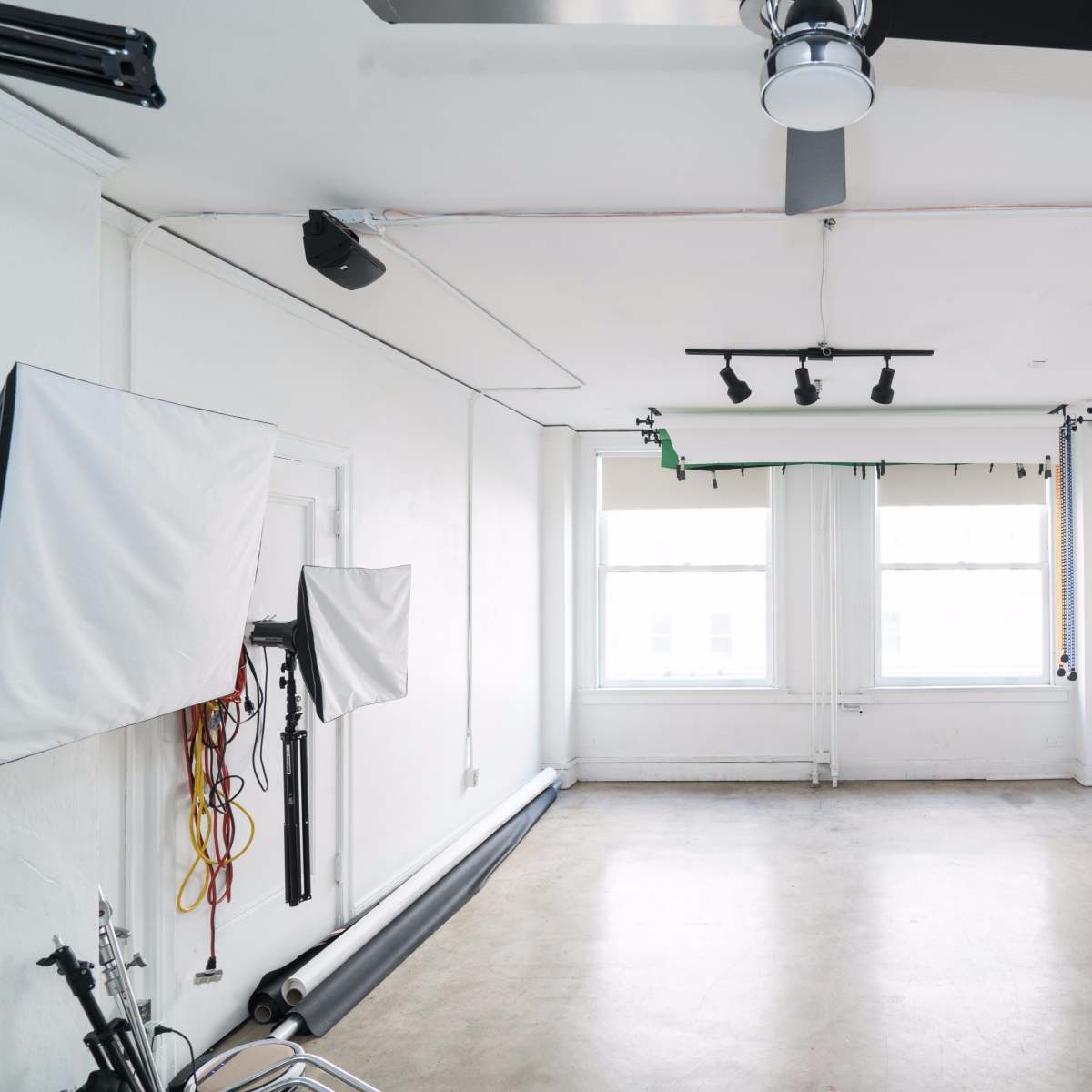 A spacious, empty studio with concrete flooring, white walls, two large windows, and a setup for photography that includes light stands and a folding table with chairs.