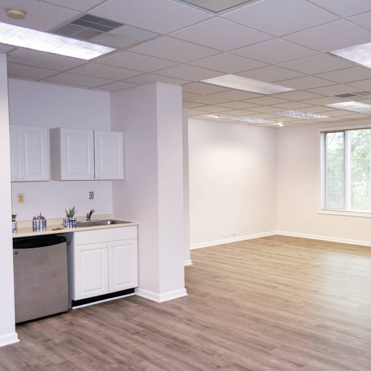 An empty, well-lit room with wooden flooring, a kitchen area featuring a small refrigerator and white cabinets, and large windows providing natural light.