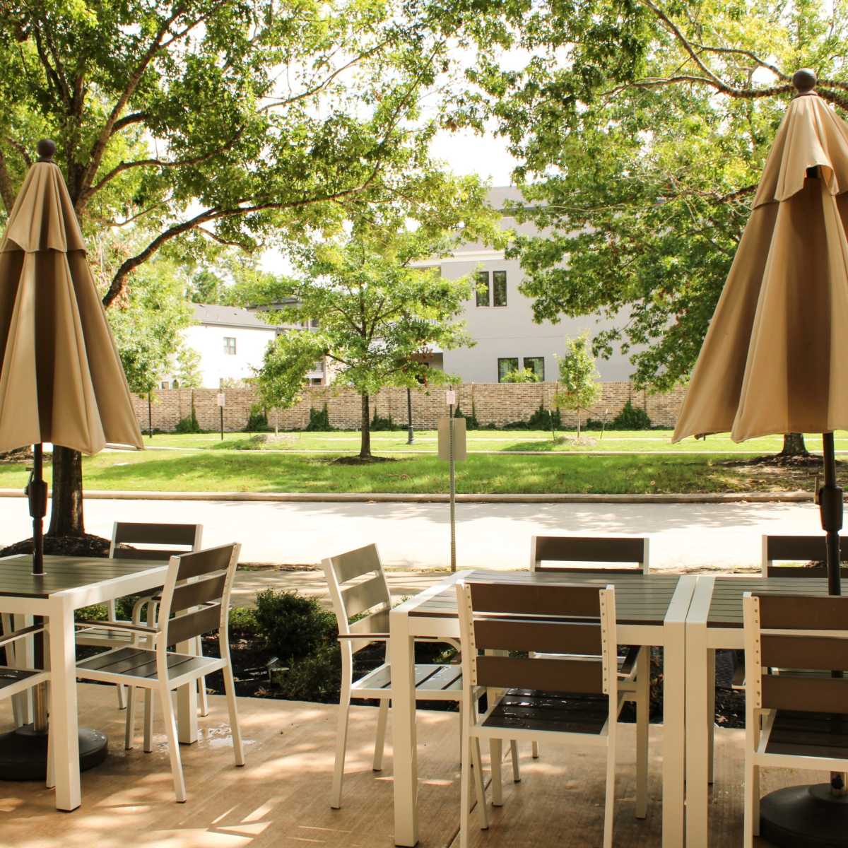 An outdoor seating area with white tables and brown chairs is shaded by large umbrellas, overlooking a tree-lined street.