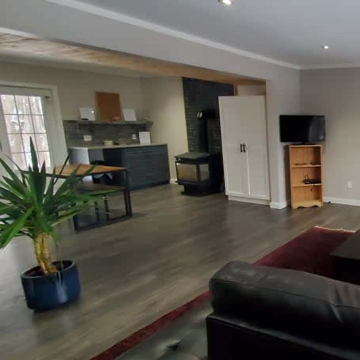 A living room with a dark leather sofa, a coffee table, a floor lamp, and large windows letting in natural light, adjacent to a kitchen area.