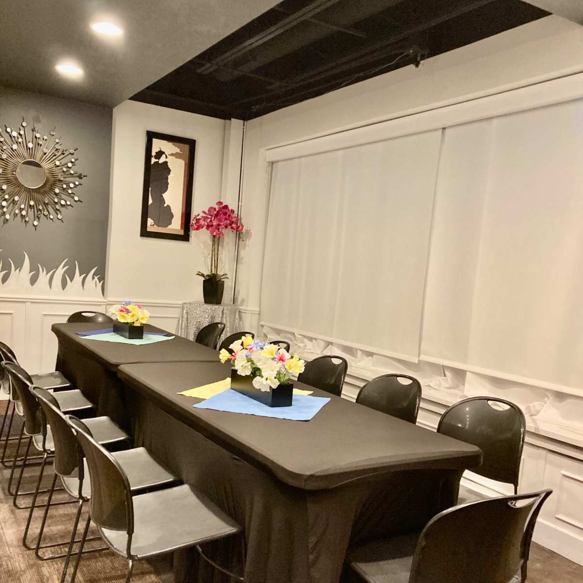 A long black table is set with flower centerpieces in a neatly arranged meeting room with black chairs and soft lighting.