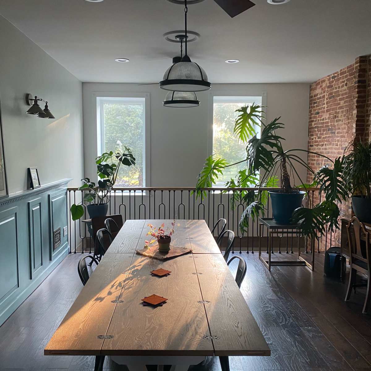 A spacious dining area features a long wooden table surrounded by chairs, with large windows and various indoor plants accentuating the brick and paneled walls.