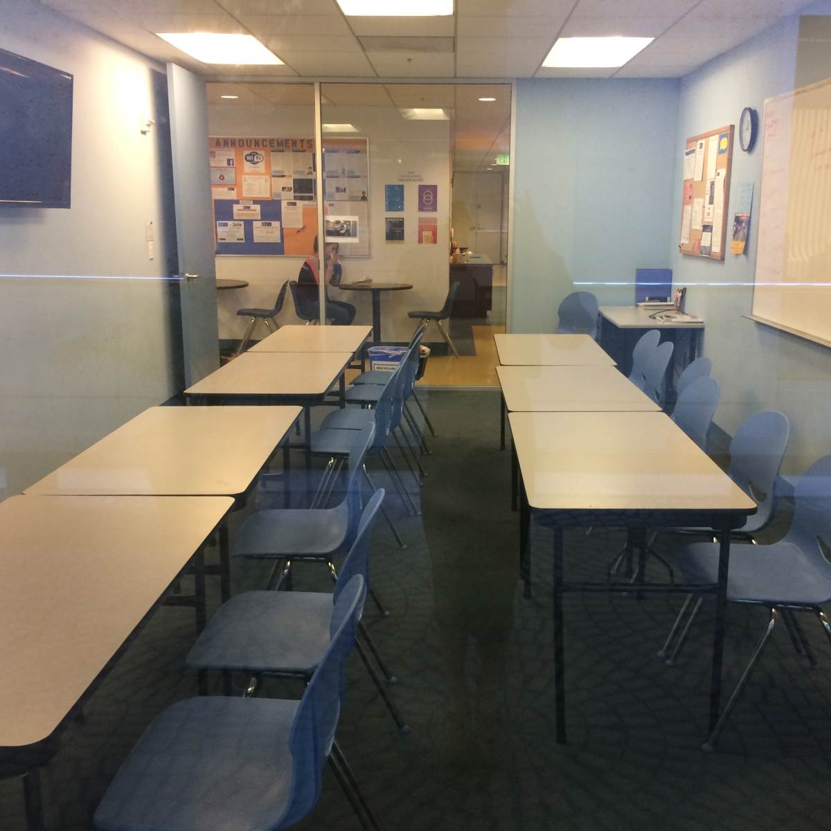 A classroom with several tables arranged in a U-shape and blue chairs, visible through glass walls, with educational materials on the walls.