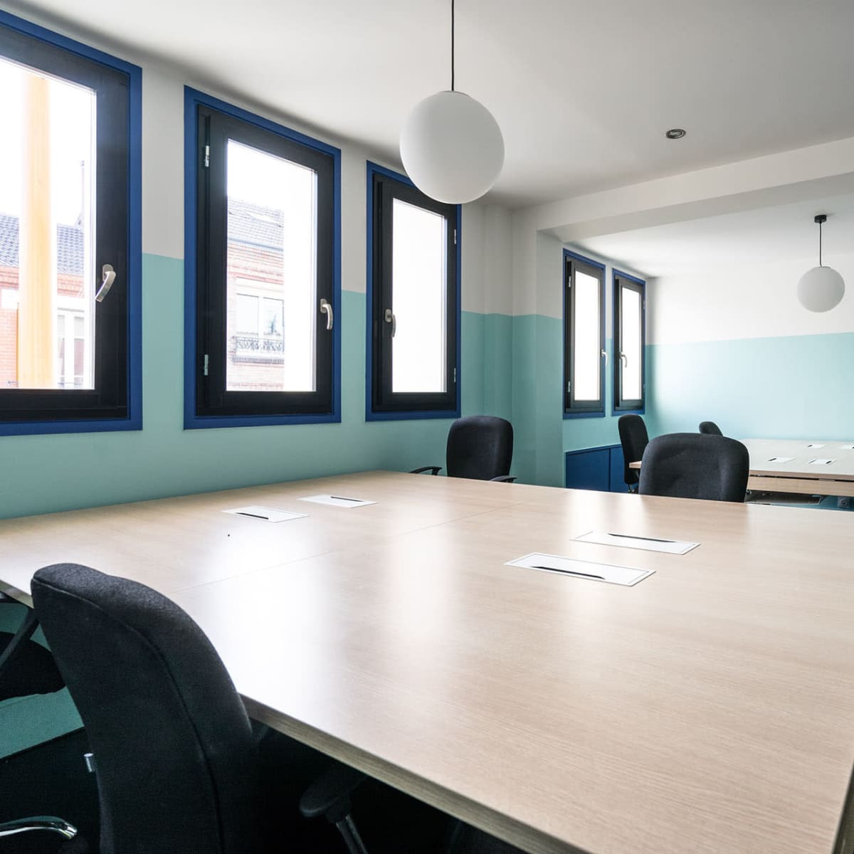 A modern conference room features several large tables with ergonomic chairs, blue and white walls, and multiple windows allowing natural light.