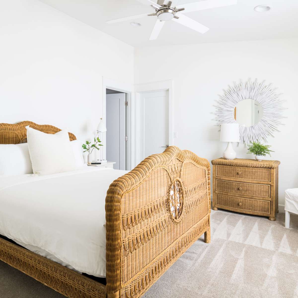 A bright bedroom featuring a rattan bed frame, a nightstand with a lamp, a dresser, and a chair, all set against white walls and light-colored flooring.