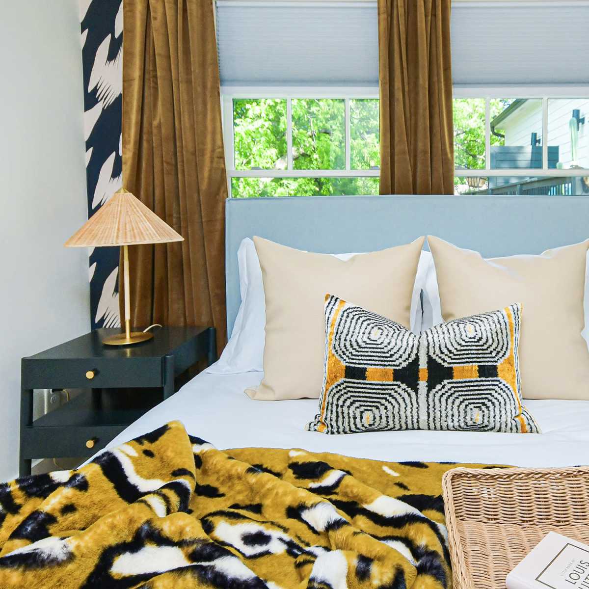 A neatly made bed with decorative pillows is positioned between two bedside tables, adorned with lamps and a small plant, against a backdrop of large windows with patterned curtains.