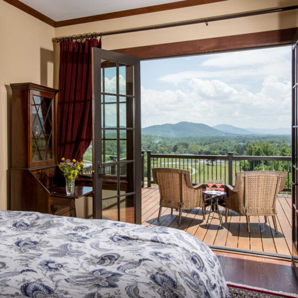 A spacious bedroom features a bed with patterned bedding, large windows with red curtains, and a balcony that opens to a view of mountains and a decorative seating area.