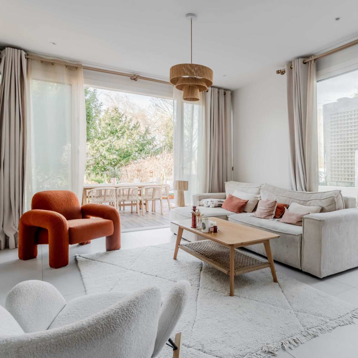 A modern living room with a white sofa, a wooden coffee table, and a red chair, featuring large windows that open to an outdoor space.