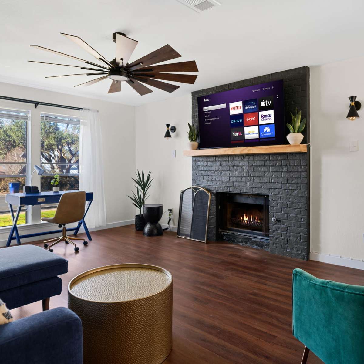 A living room features a mounted television above a black-brick fireplace, surrounded by blue and green seating, a coffee table, and a desk near a window.