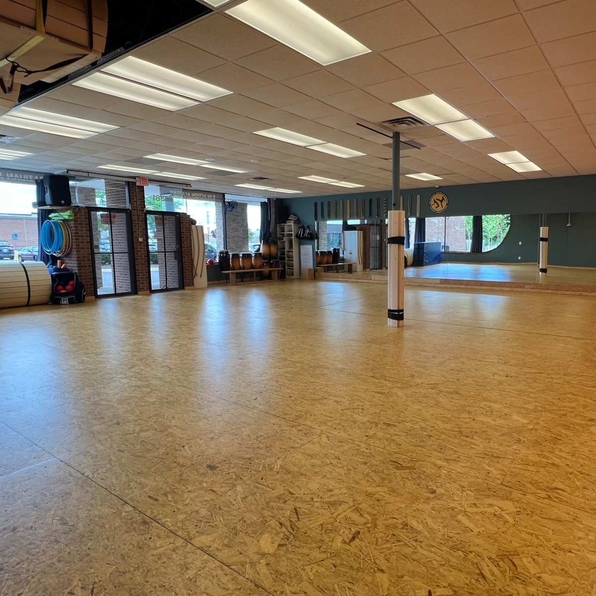 An empty indoor space with wooden flooring and orange walls, featuring various exercise equipment and large windows letting in natural light.