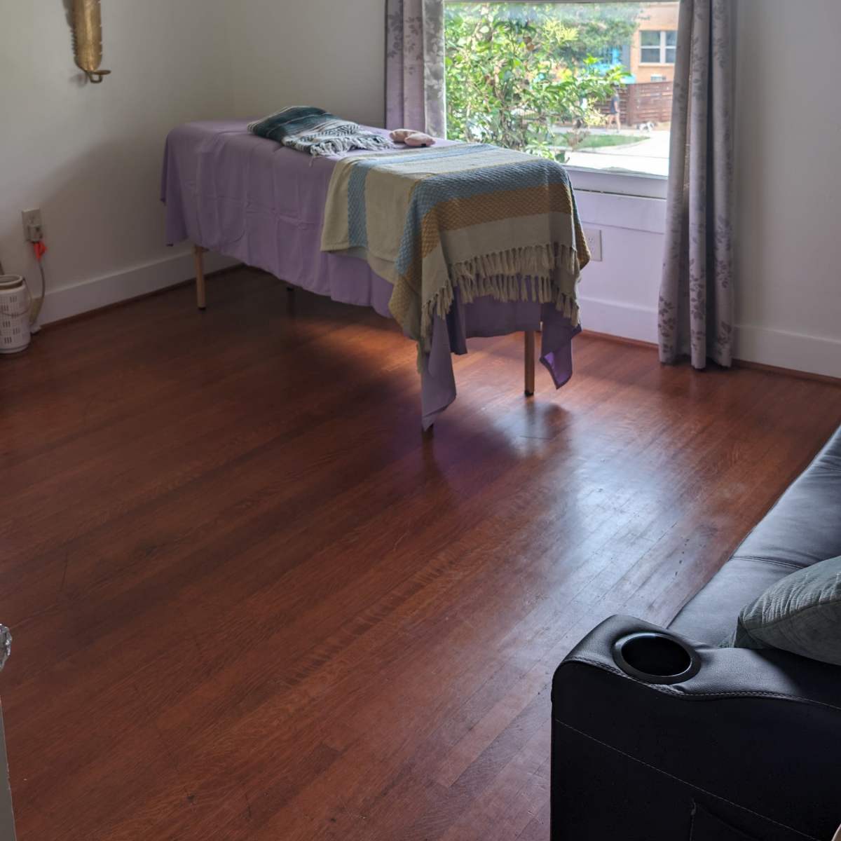 A room with wooden flooring features a table covered with a purple cloth near a window with gray curtains, and a sofa in the corner.