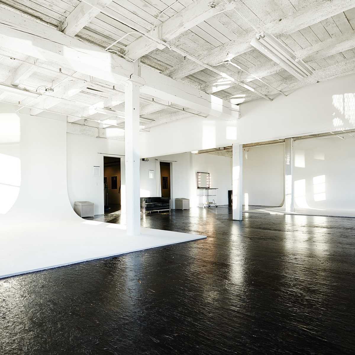 A spacious, well-lit studio with white walls, a light-colored floor, and large windows casting shadows on the surfaces.