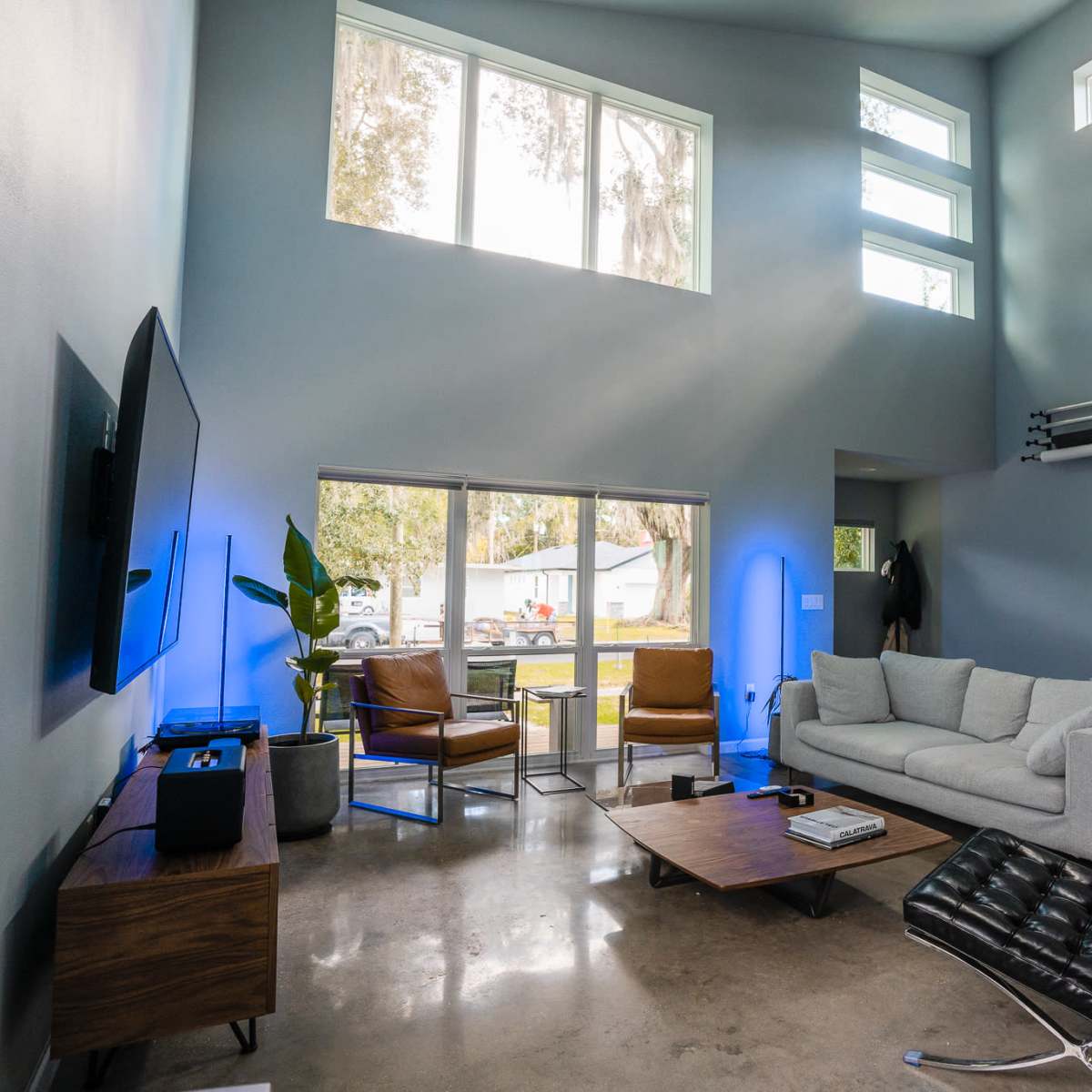 A modern living room with a gray sofa, a wooden coffee table, a television mounted on the wall, and large windows letting in natural light.