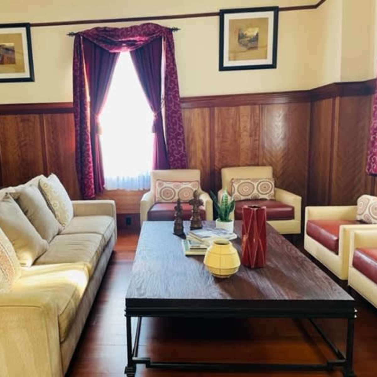 A living room with a beige sofa, a dark wooden coffee table, and two red and cream armchairs, all set against wooden-paneled walls and decorated with framed artwork.