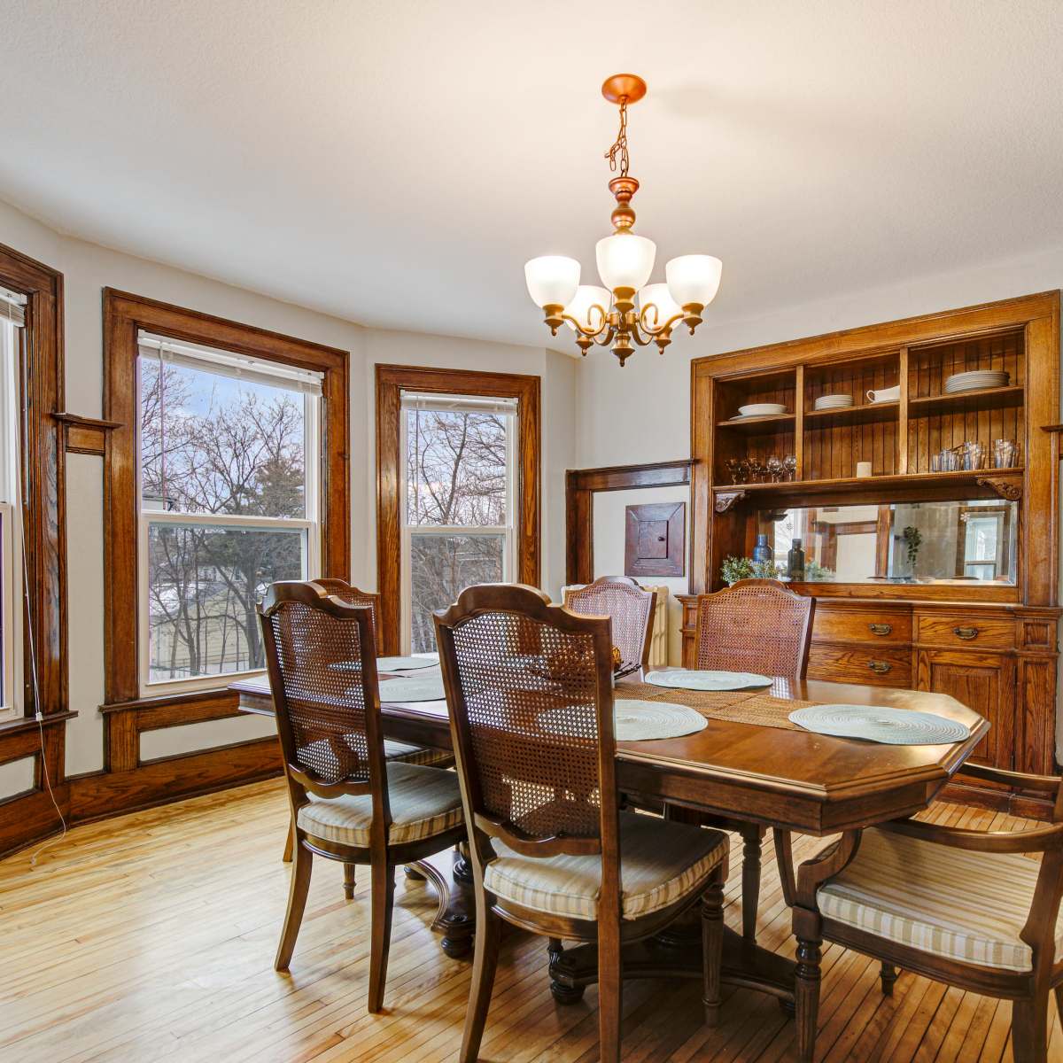 A dining room features a wooden table with six chairs, a chandelier overhead, and large windows with views of the outdoors.