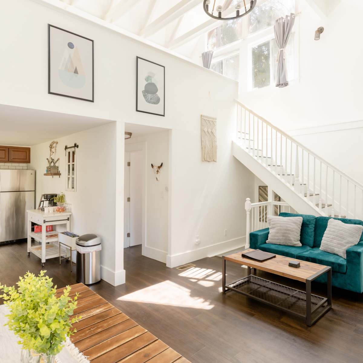 A bright, modern living space featuring a staircase, a teal sofa, and a dining table with a vase of flowers.