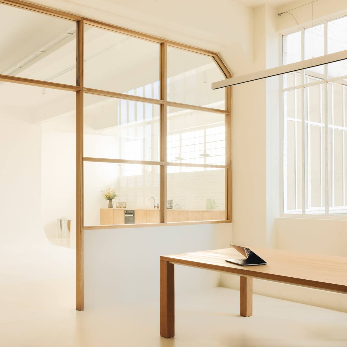 A minimalist interior features a large wooden table in the foreground and a glass partition separating a bright kitchen area in the background.