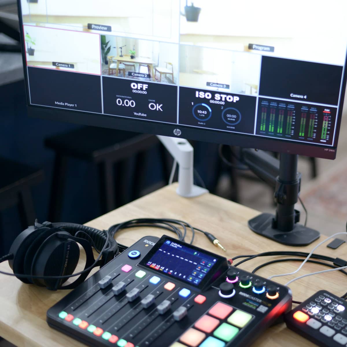 A computer monitor displays multiple camera angles while a audio mixer, a microphone, and headphones are arranged on a wooden desk.