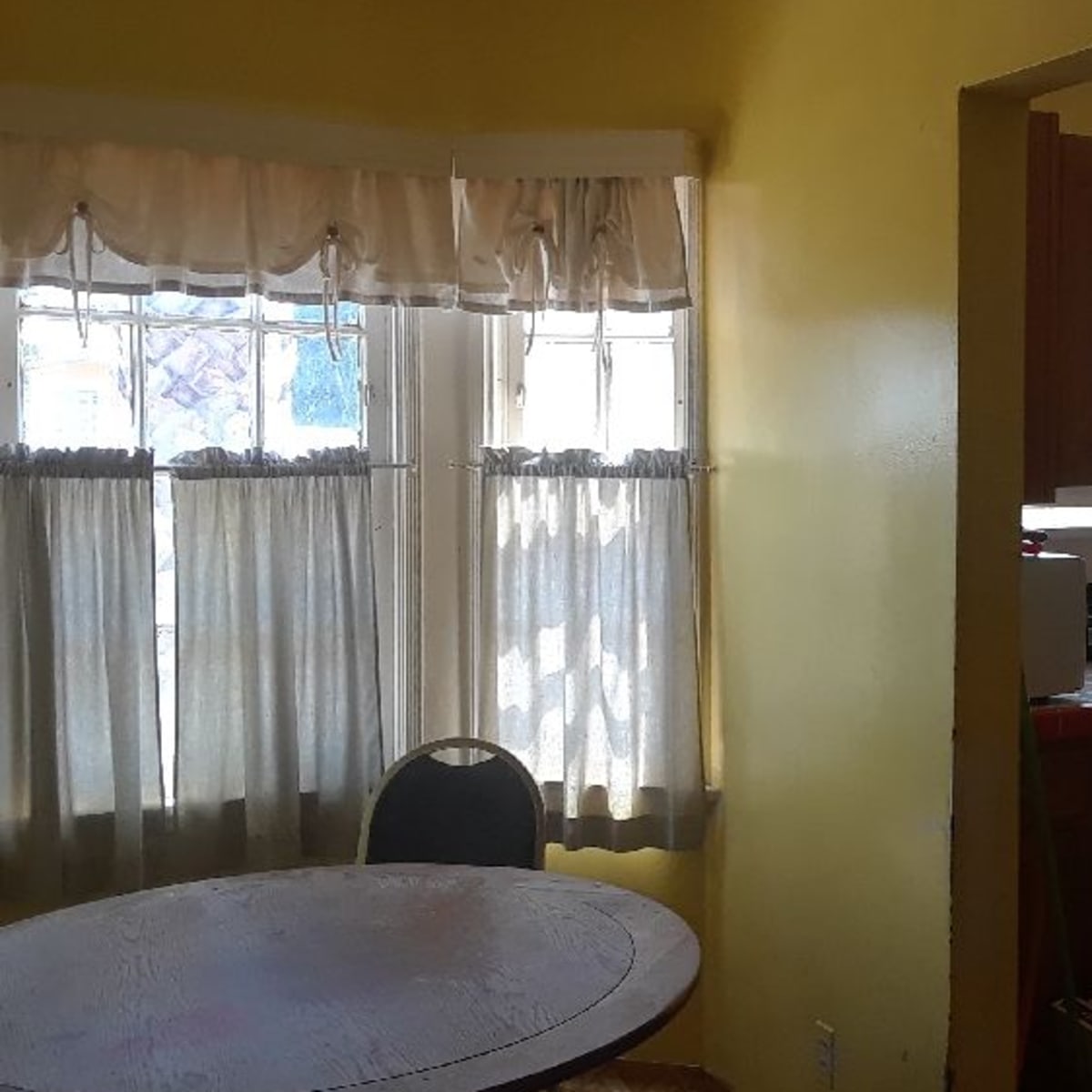 A small kitchen nook features a round table and large windows with sheer curtains, all set against yellow walls.