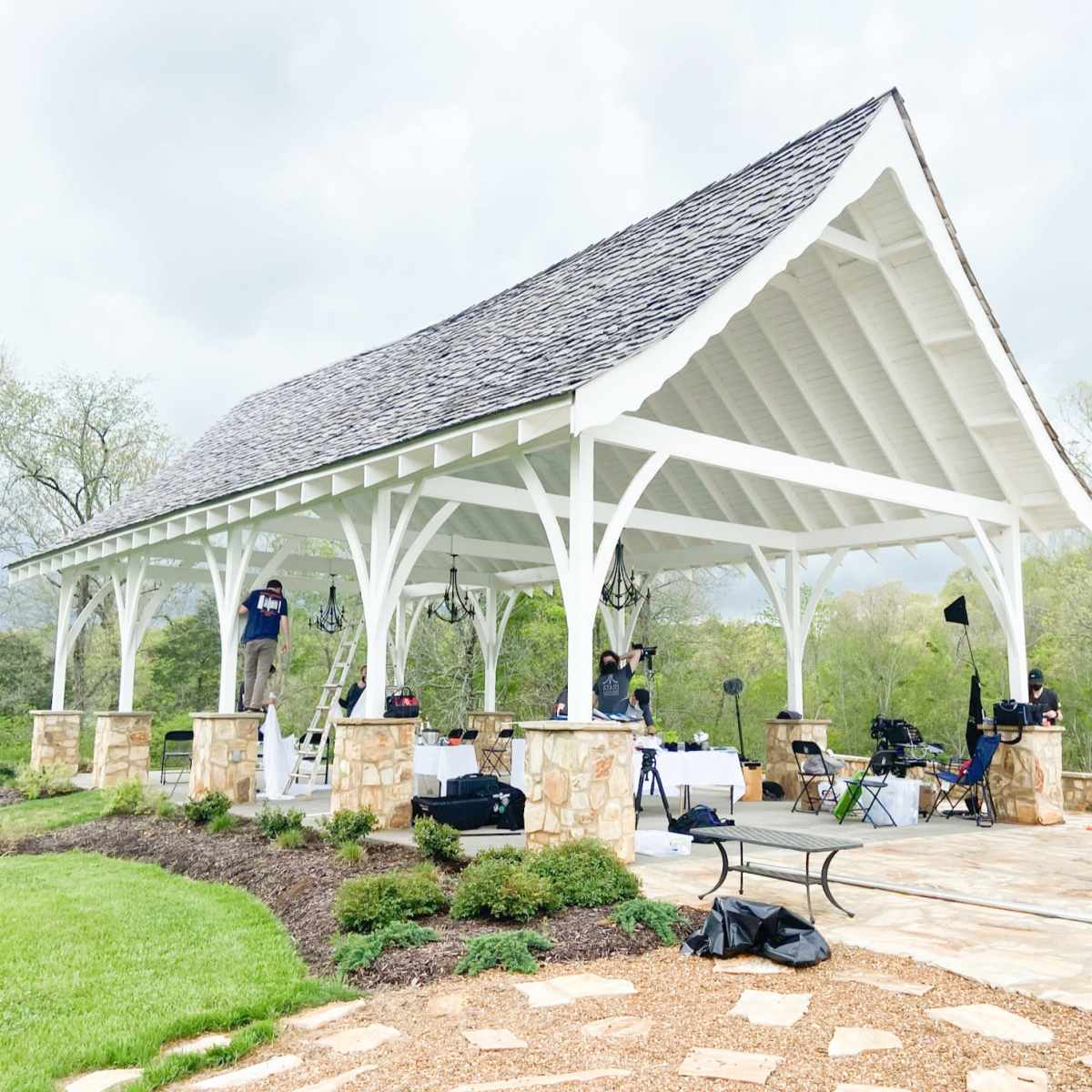 A large white pavilion with a slanted roof is set in a green outdoor area, where several people are working on camera equipment.