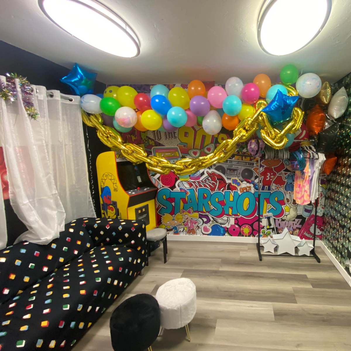A vibrant party room adorned with colorful balloons, a large banner reading "STARSHOTS," and various decorations on the walls, including sequined panels and a neon rainbow light.