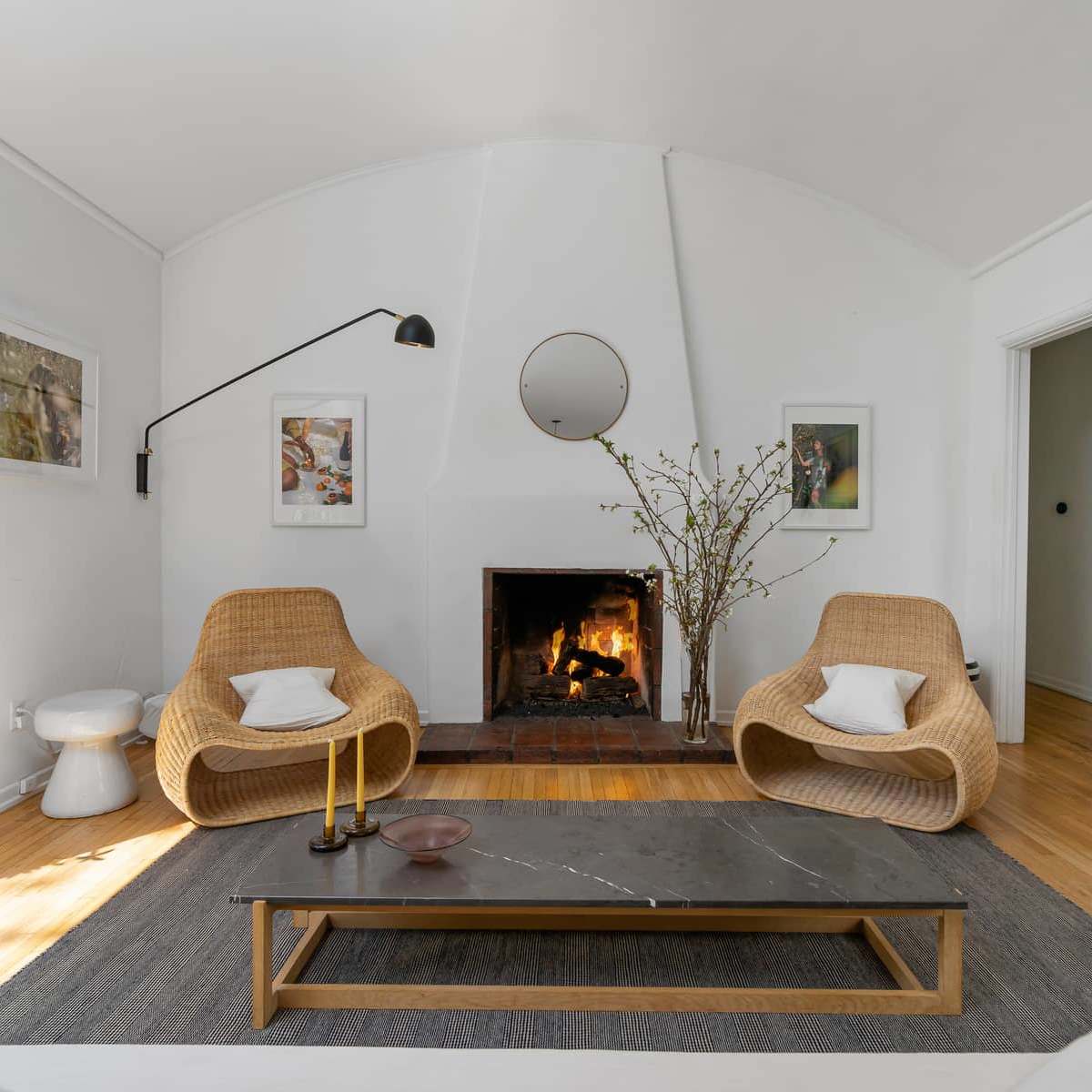A living room features two rattan chairs facing a coffee table over a rug, with a fireplace and framed pictures on the white walls.
