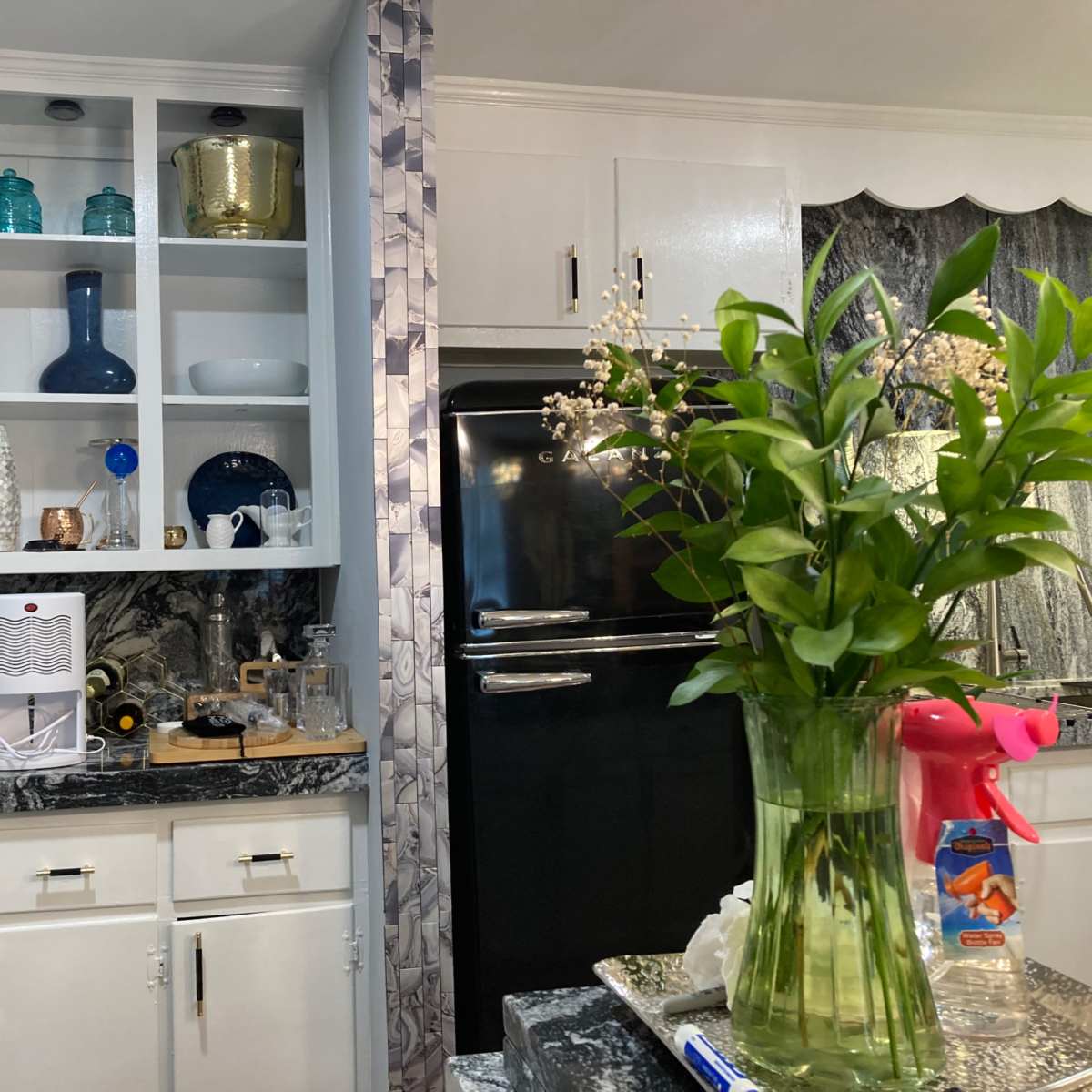A kitchen with upper shelves displaying decorative items and tableware, alongside a black refrigerator and a counter with cleaning supplies and a vase of flowers.