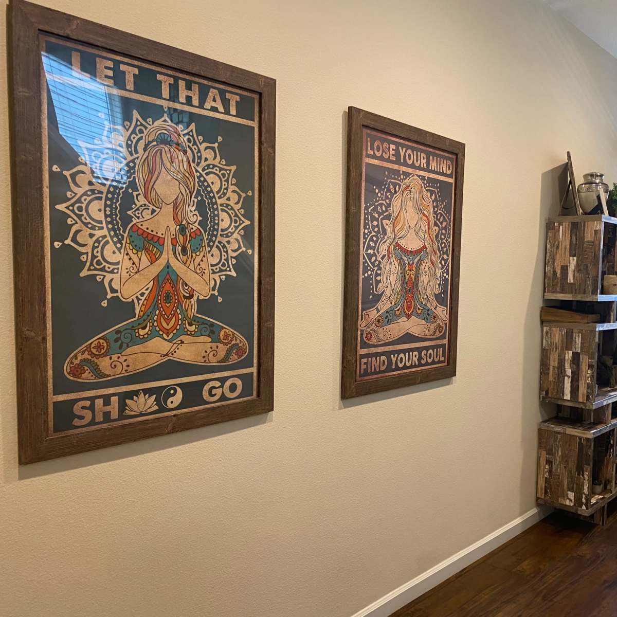 A hallway with two framed wall art pieces featuring meditative figures, and a wooden shelving unit displaying plants and decorative items.
