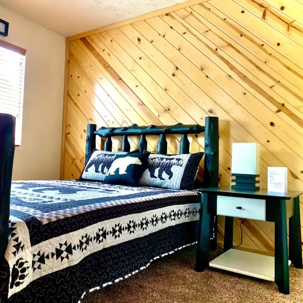 A cozy bedroom featuring a wooden bed with a black and white patterned quilt, a nightstand, and a wall with wooden paneling.