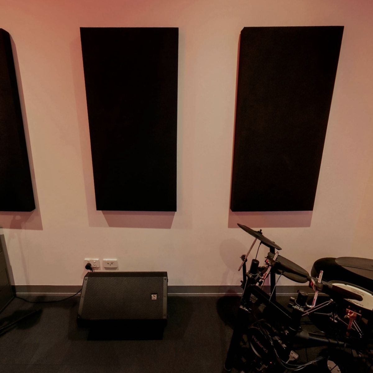 A music studio corner with three black acoustic panels on the wall, a microphone, an electronic drum set, and a speaker positioned on the floor.