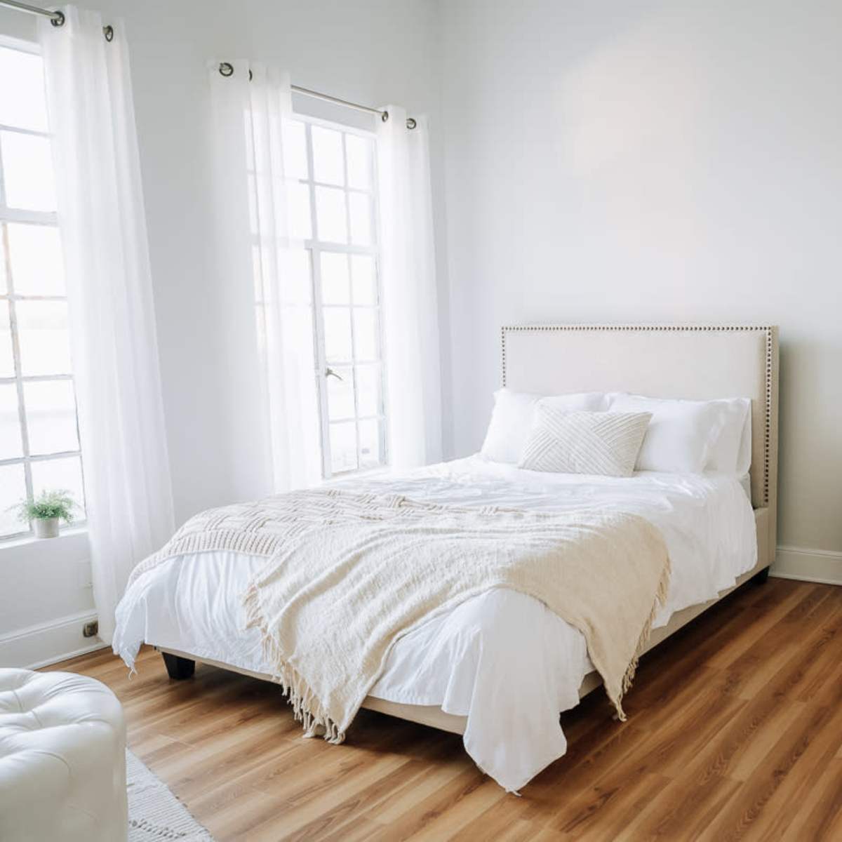 A light-colored bedroom featuring a bed with a beige throw, a wooden dresser, and large windows with sheer curtains.