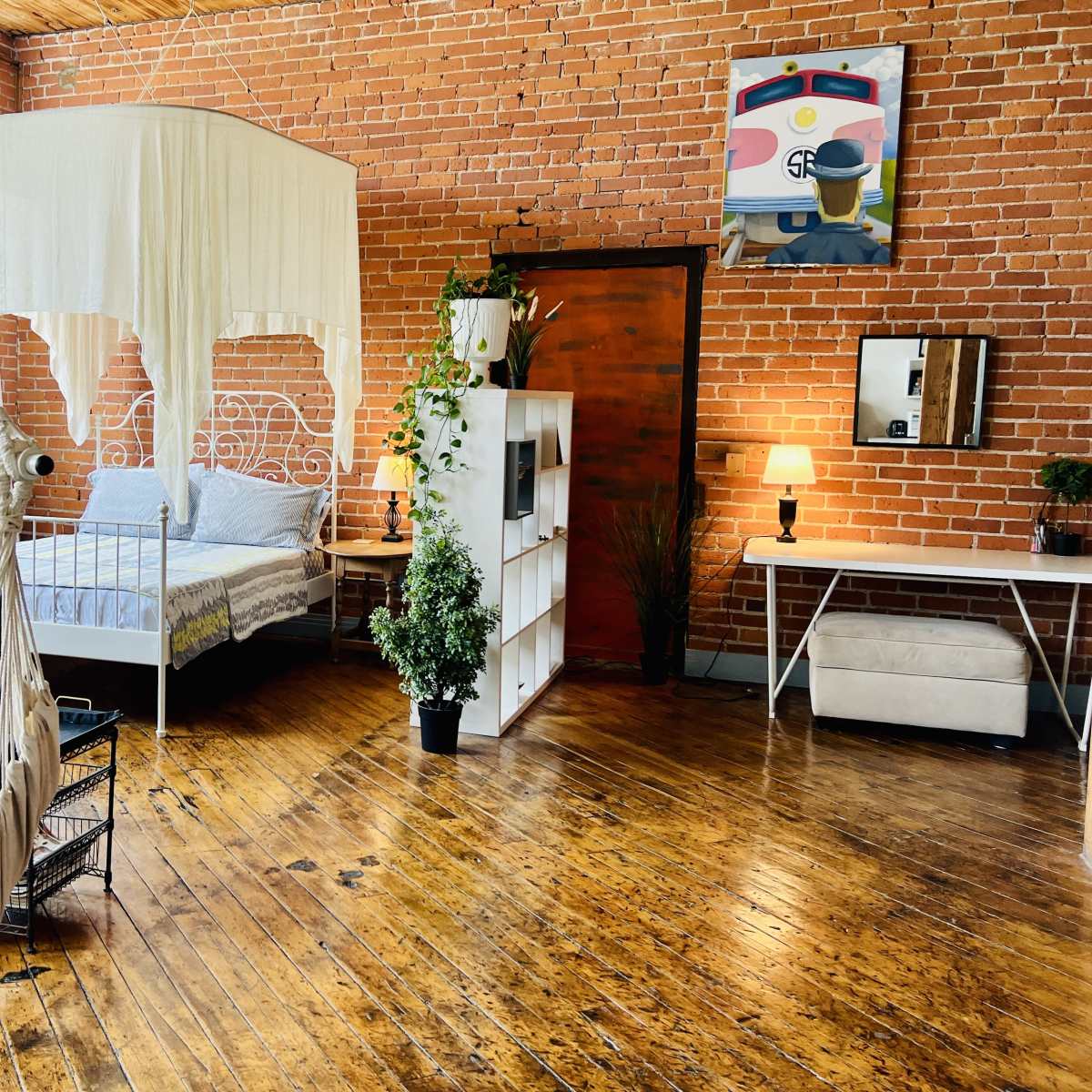A spacious room with exposed brick walls featuring a bed with a canopy, a hammock chair, a white shelving unit, and various plants.