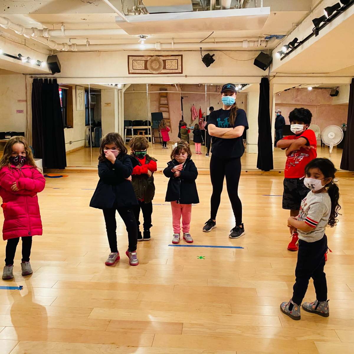 A group of six children and one adult wearing masks stands in a dance studio with wooden floors and mirrors.