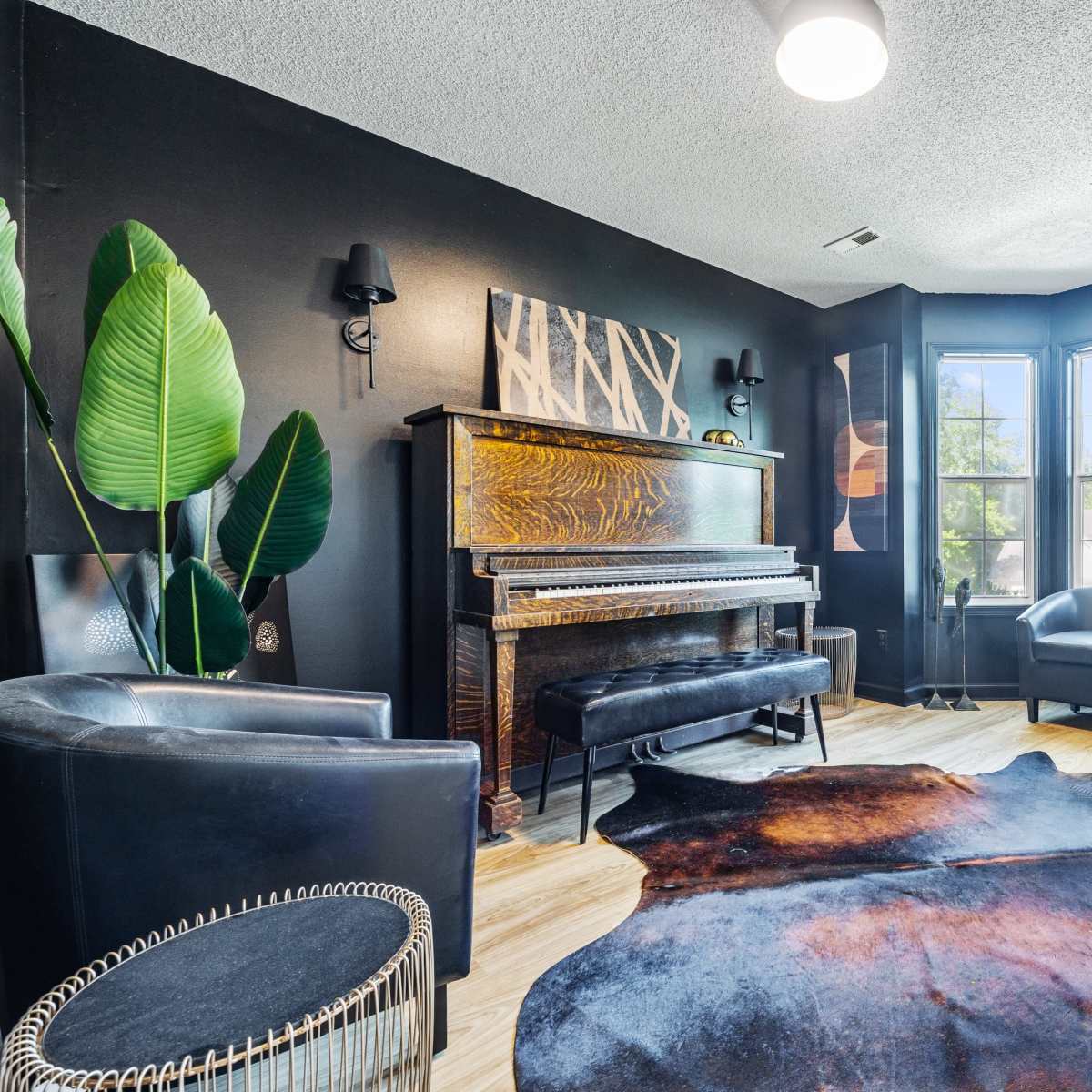 A corner of a room features a piano, two black chairs, a cowhide rug, and large windows with decorative wall art.