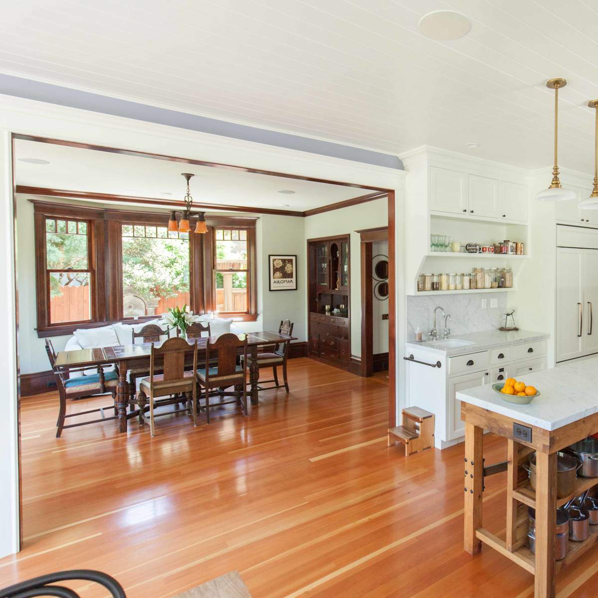 A spacious kitchen features a central island with a marble top and wooden base, leading to an adjoining dining area with a large table and chairs.