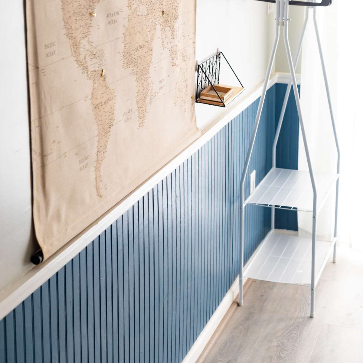 A wall with a world map tapestry, a geometric decorative item on a ledge, and a simple white clothing rack in a room with blue wainscoting.