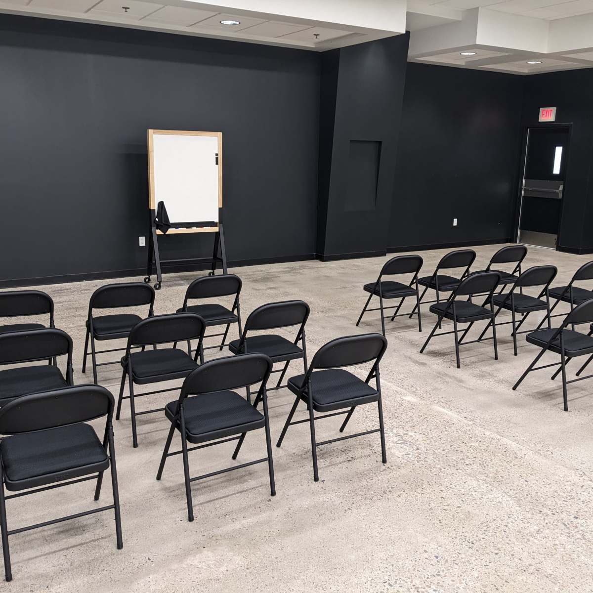 A row of black folding chairs faces a whiteboard in a minimalist, empty room with dark walls and a concrete floor.